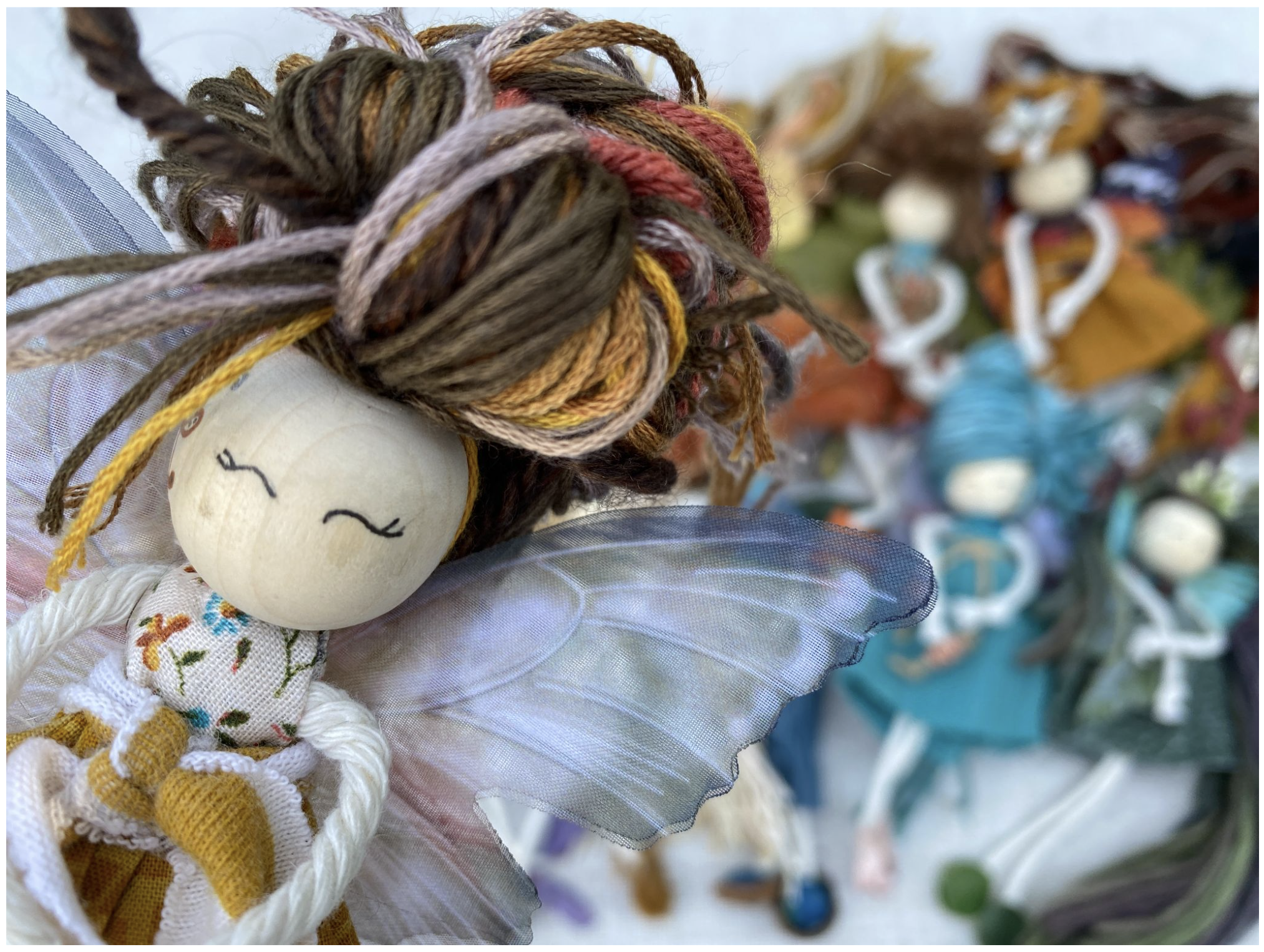 Close-up of a handmade fabric faerie doll with yarn hair and tulle wings, with more faeries blurred in the background